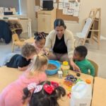 A teacher and several young children gather around a table, mixing ingredients in a large blue bowl. Various containers, including one of milk, are on the table, as the children watch and participate in the activity.