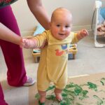 A smiling baby in a yellow romper stands on a floor covered with green paint, supported by an adult holding their arms. The baby’s feet are painted green, and art supplies are visible in the background.