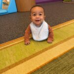 A smiling baby wearing a striped bib and brown long-sleeve top is crawling on a colorful carpet in a playroom with purple walls and various play structures in the background.
