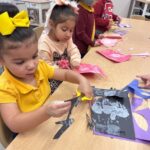 Four young children sit at a classroom table, focused on cutting and gluing colored paper for an art project. The table is filled with craft supplies, and the kids appear engaged and creative.