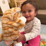 A smiling toddler with dark hair, wearing a white shirt and pink pants, plays with a plush orange-and-white striped cat puppet in a cozy indoor setting.