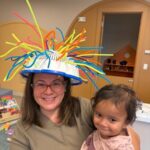 A woman wearing glasses and a helmet decorated with colorful pipe cleaners smiles while holding a young child in a pink outfit. They are indoors, with toys and shelves visible in the background.