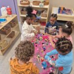 Five young children play together at a sensory table filled with colorful pom-poms and scoops in a classroom. Shelves with baskets and learning materials are visible in the background.