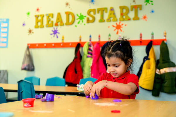 A young child in a classroom shapes purple playdough at a table. Colorful coats hang on hooks behind her, and HEAD START is displayed on the wall with bright decorations.