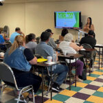A woman stands at the front of a classroom giving a presentation to seated adults. A screen displays a green slide titled Mi Casa with text below. The room has colorful tiled flooring and desks arranged in rows.