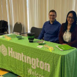 Two people sit behind a green table with a Huntington logo, smiling at the camera. The table has brochures, small items, and a cup of pens. They are in a room with chairs and vertical blinds in the background.