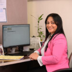 A woman in a pink blazer sits at a desk in an office, smiling at the camera. A computer monitor, documents, a plant, and office supplies are visible on the desk.