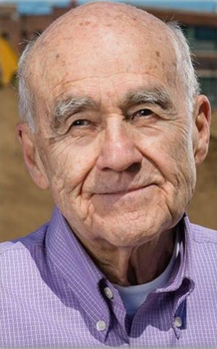 An elderly man with a bald head and gray eyebrows, wearing a light purple button-up shirt, looks at the camera with a gentle expression. The background is blurred, showing some outdoor elements.