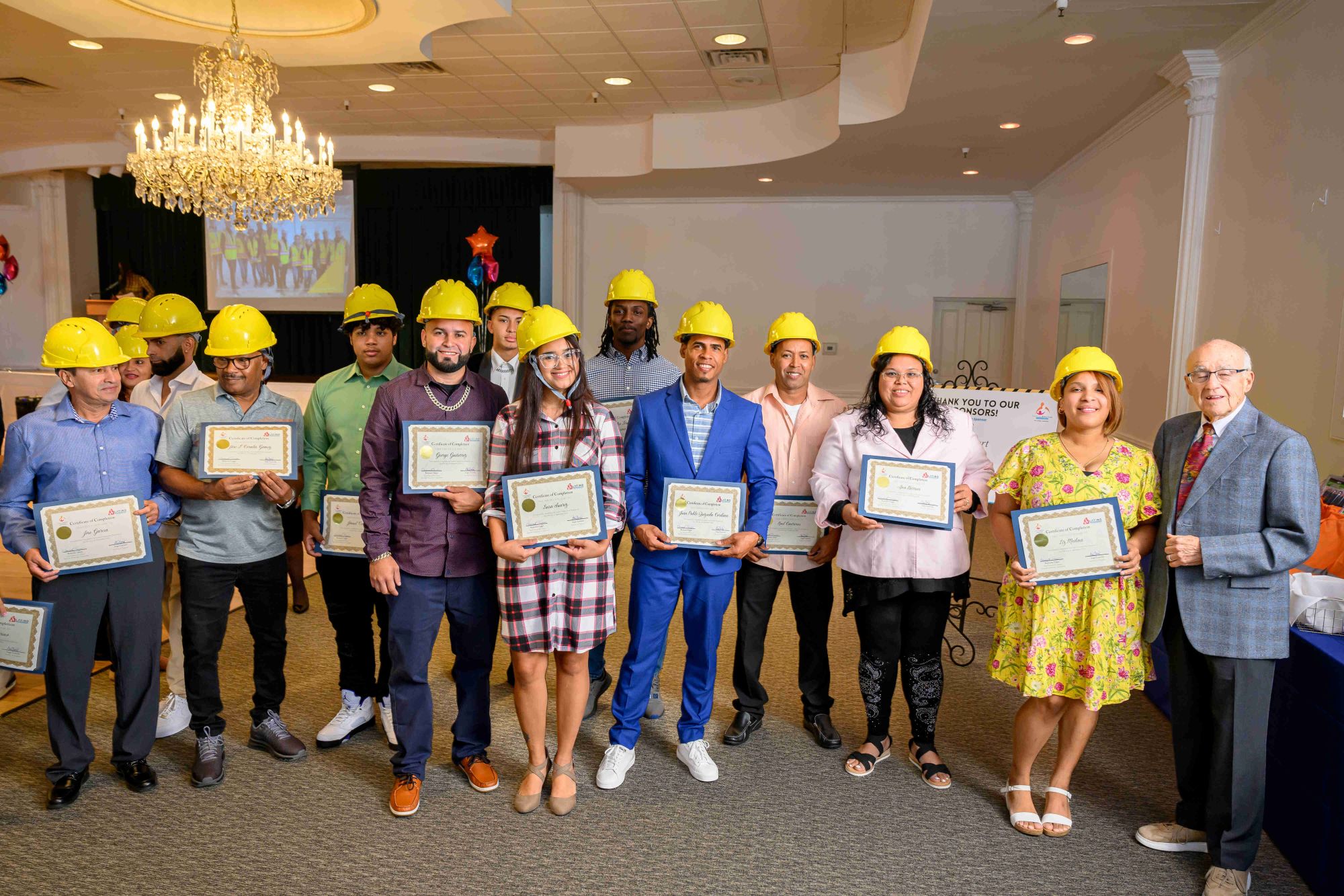 A group of people wearing yellow construction helmets stand indoors, holding certificates and smiling at the camera. An older man in a suit stands at the far right. The setting appears to be a celebratory event or graduation.