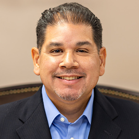 Smiling man with short dark hair and goatee, wearing a blue collared shirt and dark blazer, seated against a neutral background.