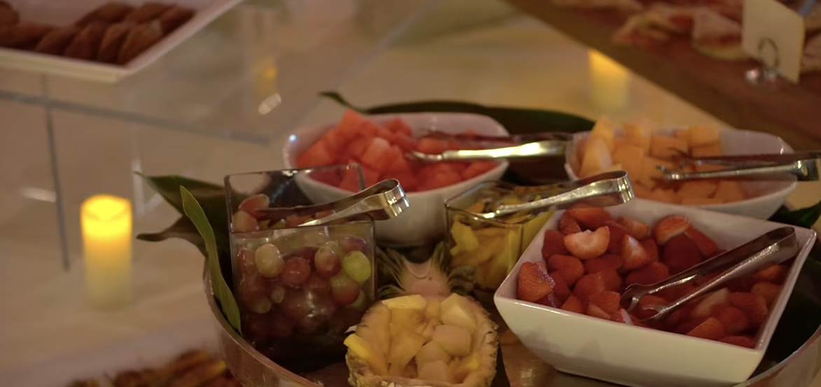 A table display with bowls of assorted fresh fruits, including grapes, strawberries, cantaloupe, and watermelon, arranged with serving tongs. A lit candle glows softly in the background.