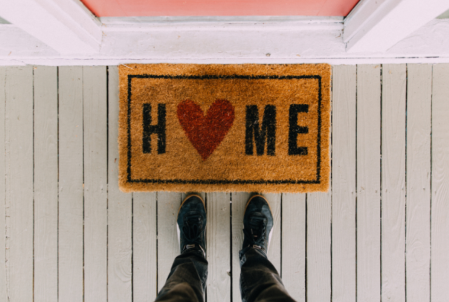 A person in black shoes stands on a wooden porch in front of a doormat that says HOME, with a red heart symbol replacing the letter O.