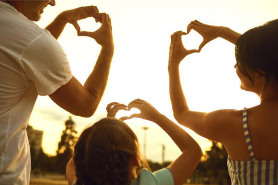 A family of three faces the sunset, holding up their hands to form heart shapes together. The warm light creates a joyful and loving atmosphere.