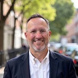 A man with short hair, glasses, and a beard, wearing a white shirt and dark blazer, smiles outdoors on a city street with trees and buildings in the background.