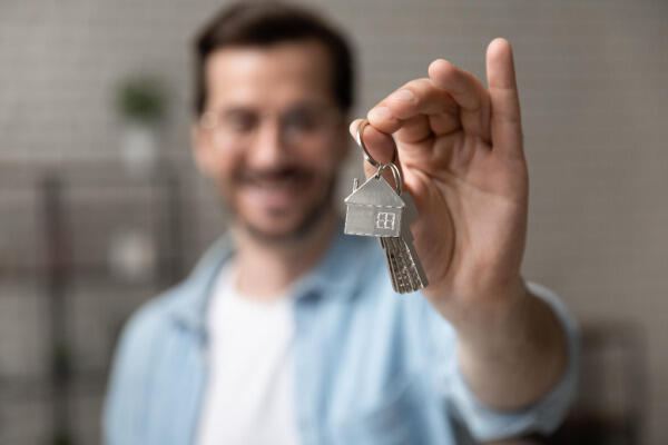 A smiling man holds up a set of house keys with a house-shaped keychain, focusing on the keys in the foreground while the man is blurred in the background.