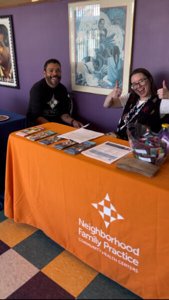 Two people sit behind an orange table with Neighborhood Family Practice Community Health Centers on the front. The table has brochures and items, and both people are smiling. Art and colorful floor tiles are visible.
