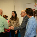 A group of people stand indoors, smiling and shaking hands. One person holds a framed certificate. A banner in the background reads “Spanish American” and shows a logo with three figures.