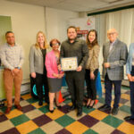 Eight people stand indoors on a colorful tiled floor. One person in the center holds a framed certificate. They are all dressed in business or semi-formal attire, smiling and facing the camera.