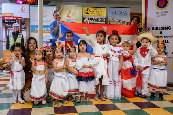 A group of young children and two adults in traditional and festive clothing pose together indoors, some holding a large flag. Colorful decorations and banners are visible in the background.