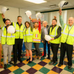 A group of eight people wearing safety vests and holding hard hats stand indoors on a colorful tiled floor, smiling and raising their arms in celebration.