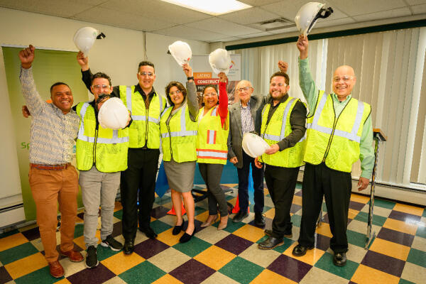 A group of eight people wearing safety vests and holding hard hats stand indoors on a colorful tiled floor, smiling and raising their arms in celebration.