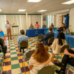 A man stands and speaks at the front of a room to a group of seated people. The audience sits around tables covered with blue cloths, and the floor features a colorful checkered pattern.