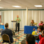 A man speaks at a podium in a room with seated attendees. Three people sit at a blue-draped table facing the audience. Certificates and a safety helmet are visible on display to the side.