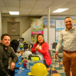 Three people, two sitting at a table and one standing, smile at the camera in a meeting room. The table has hard hats, water bottles, and papers. Colorful banners and empty chairs are in the background.