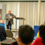 An older man speaks at a podium to an audience in a conference room. People sit at tables, listening. A sign reads Latino Construction Program. One woman watches the speaker attentively.