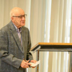 An elderly man wearing glasses and a patterned gray blazer stands beside a lectern, holding a piece of paper and pen, speaking in front of vertical blinds.