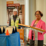 A woman in a pink blazer stands at a podium speaking. Next to her, a table holds several certificates. Behind her is a banner for the Latino Construction Program featuring a smiling worker in safety gear.