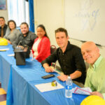Six people sit at a long table covered with a blue cloth, smiling at the camera. Construction helmets and water bottles are on the table. Behind them, a whiteboard reads LCP class of XV.