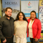 Three people stand smiling in front of colorful banners promoting Spanish American Committee programs, including financial literacy and family services. Two women are on the right and a man is on the left.
