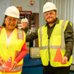 Two people wearing hard hats, safety glasses, and yellow vests smile while holding small orange traffic cones. A certificate and a construction program sign are visible in the background.