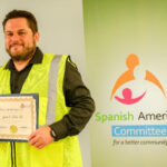 A man wearing a yellow safety vest holds a certificate and stands in front of a banner that reads Spanish American Committee for a better community. He is smiling and posing for the photo.