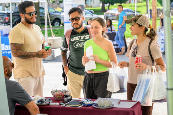 Four people stand at an outdoor table, talking to a seated person. Two women hold shopping bags, one with a can of soda, while two men stand nearby. Informational booths are visible in the background.