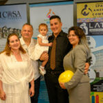 Five people pose and smile in front of banners for community organizations. A man holds a toddler in a white dress; another woman in a suit holds a yellow hard hat. All are dressed formally, suggesting a special occasion or event.