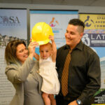A woman and man smile as they help a toddler wear a yellow construction helmet. They stand indoors in front of banners for housing and construction programs, including Mi Casa and Spanish American Committee.