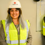 A person wearing a white hard hat labeled Ozanne and a yellow safety vest smiles at the camera. Another person in a safety vest stands in the background near a whiteboard.