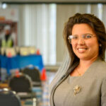 A woman with glasses and long hair, wearing a gray blazer with a brooch, smiles at the camera in a brightly lit conference room with empty chairs and blurred background.