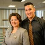 A smiling woman in a gray suit and glasses stands next to a smiling man in a black shirt and brown striped tie. They are indoors with chairs and people in the background.