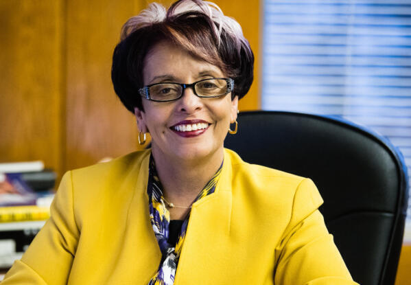 A smiling woman with short dark hair and glasses sits at a desk, wearing a bright yellow blazer and patterned scarf. There are books and a window with blinds in the background.