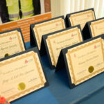 A close-up view of several certificates of completion displayed on a blue tablecloth, each mounted in black frames with gold seals, ready for presentation at an event.