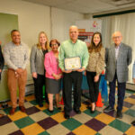 Eight professionally dressed adults stand in a row indoors; one man in the center holds a framed certificate. They smile at the camera, and colorful signs and curtains are visible in the background.