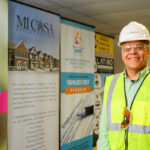 A man wearing a hard hat and safety vest smiles while standing indoors in front of banners promoting community programs.