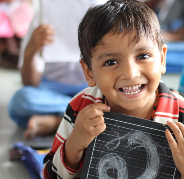 A young boy with dark hair smiles brightly while holding a small chalkboard with a drawing on it. He is wearing a red and white striped shirt and appears to be in a classroom with other children in the background.