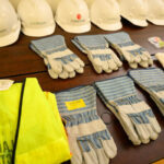 A table displays white construction helmets, high-visibility safety vests, blue and gray work gloves, red and white gloves, and earplugs in plastic bags arranged in neat rows.