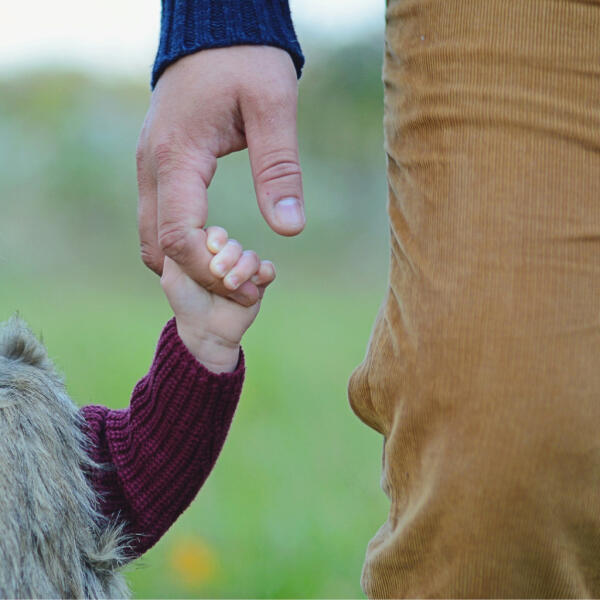 Close-up of an adult and a child holding hands outdoors. The adult wears brown pants and a blue sweater, while the child wears a maroon sweater and furry vest. The background is blurred greenery.