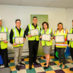 Six people wearing safety vests stand indoors, smiling and holding certificates. One person wears a white hard hat and another wears a reflective vest. They appear to be celebrating a construction-related achievement.
