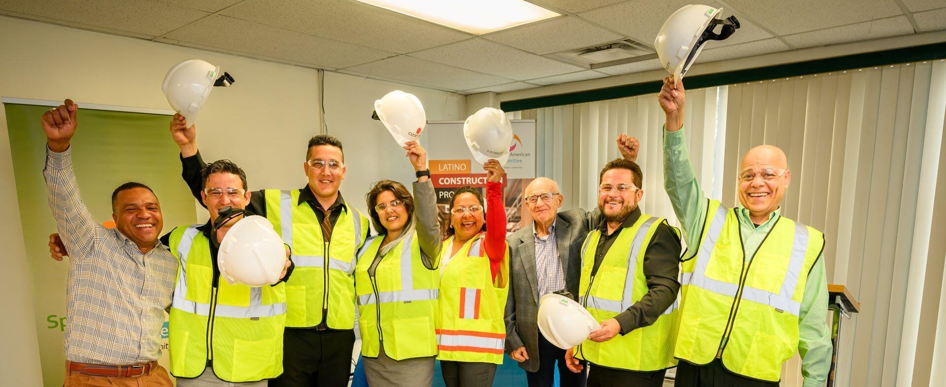 A group of eight people wearing yellow safety vests smile and raise safety helmets in the air, celebrating together in an indoor setting. A Latino Construction sign is visible in the background.